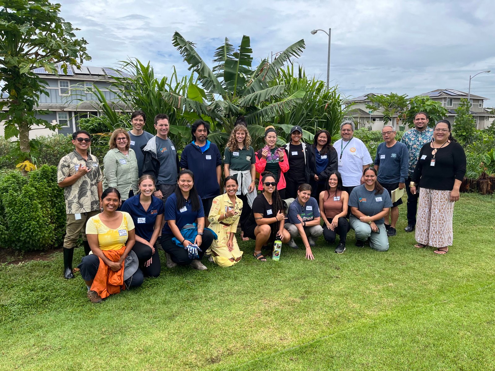 Group photo of hui members who attended the in-person convening in May of 2024. The group is standing in front of the Council for Native Hawaiian Advancement garden. A banana tree, papaya trees, cane sugar and other plants are in the background, and the members are smiling and throwing shakas in the foreground.